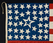 39 STARS IN A LINEAL PATTERN WITH A HUGE, HALOED CENTER STAR AND DYNAMIC VISUAL FEATURES, ITS CANTON RESTING ON THE WAR STRIPE, PROBABLY MADE FOR THE 1876 CENTENNIAL OF AMERICAN INDEPENDENCE, NEVER AN OFFICIAL STAR COUNT, REFLECTS THE ANTICIPATED ARRIVAL OF COLORADO AND THE DAKOTA TERRITORY