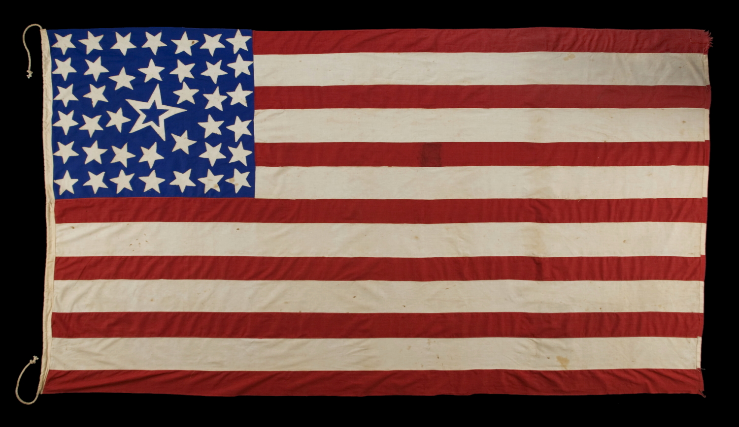 39 STARS ON A HOMEMADE, ANTIQUE AMERICAN FLAG WITH AN EXTREMELY RARE COMBINATION OF JUSTIFIED COLUMNS AND A HUGE, HALOED CENTER STAR, INFUSED AMONG THEM, WITH EXTRAORDINARY COLORS, EXCEPTIONAL VISUAL IMPACT, AND ITS CANTON RESTING ON THE WAR STRIPE; AN UNOFFICIAL STAR COUNT, REFLECT THE ANTICIPATED ARRIVAL OF COLORADO AND THE DAKOTA TERRITORY; LIKELY MADE FOR THE 1876 CENTENNIAL OF AMERICAN INDEPENDENCE