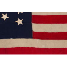 13 STAR ANTIQUE AMERICAN FLAG WITH HAND-SEWN STARS IN AN EXTREMELY NARROW PRESENTATION OF A 3-2-3-2-3 ARRANGEMENT ON A CANTON THAT DOESN’T FOLLOW SUIT, LEAVING WIDE EXPANSES OF BLUE WOOL BUNTING ABOVE AND BELOW; MADE IN THE ERA OF THE 1876 CENTENNIAL OF AMERICAN INDEPENDENCE, POSSIBLY AS A U.S. NAVY SMALL BOAT ENSIGN